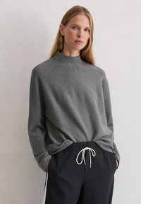 Graues Langarmoberteil mit hohem Hals, entspannter Passform, sanft strukturiertem Stoff, kombiniert mit schwarzen Jogginghosen mit Kordelzug und kontrastierendem Bund.