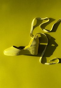 Espadrille compensée jaune avec lacets à nouer autour de la cheville, posée à plat sur un fond jaune avec des ombres marquées.
