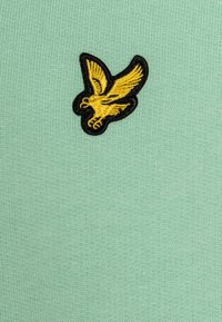 Pájaro bordado amarillo con contorno negro sobre un fondo de tela de punto verde claro.