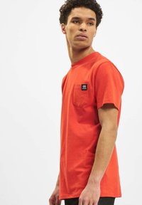 Rotes Baumwoll-T-Shirt mit kurzen Ärmeln, Rundhalsausschnitt und einer linken Brusttasche mit einem kleinen schwarzen Logo. Glatte Textur, lässiges Design.