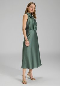 Robe en satin vert avec un col montant, taille élastique et longueur midi. Associée à des talons dorés à lanières et des bijoux assortis.