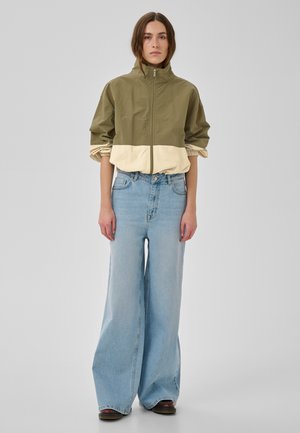 Naine, kes kannab oliivi- ja beeživärvilist lukuga jope, mille varrukaid on üles rullitud, helesiniseid laia säärega pükse ja pruunid jalatseid, seisab ühevärvilise tausta ees.