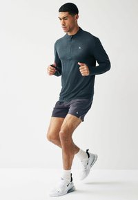Dunkelgraues Langarmshirt mit Viertelreißverschluss, kombiniert mit dunkler Shorts. Das Modell trägt weiße Sportschuhe mit schwarzen Akzenten und weiße Socken.