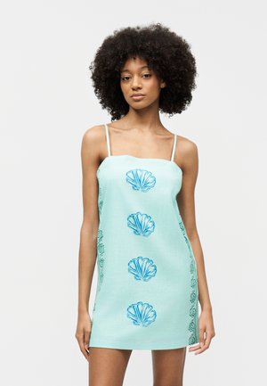 Femeie cu păr creț, purtând o rochie fără mâneci, de culoare albastru deschis, cu modele albastre de scoici și spirale, stând în fața unui fundal simplu.