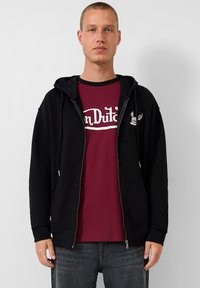 Schwarzer Zip-Hoodie mit Kängurutasche, getragen über einem burgunderfarbenen T-Shirt mit weißem Aufdruck. Das Modell trägt außerdem graue Jeans.
