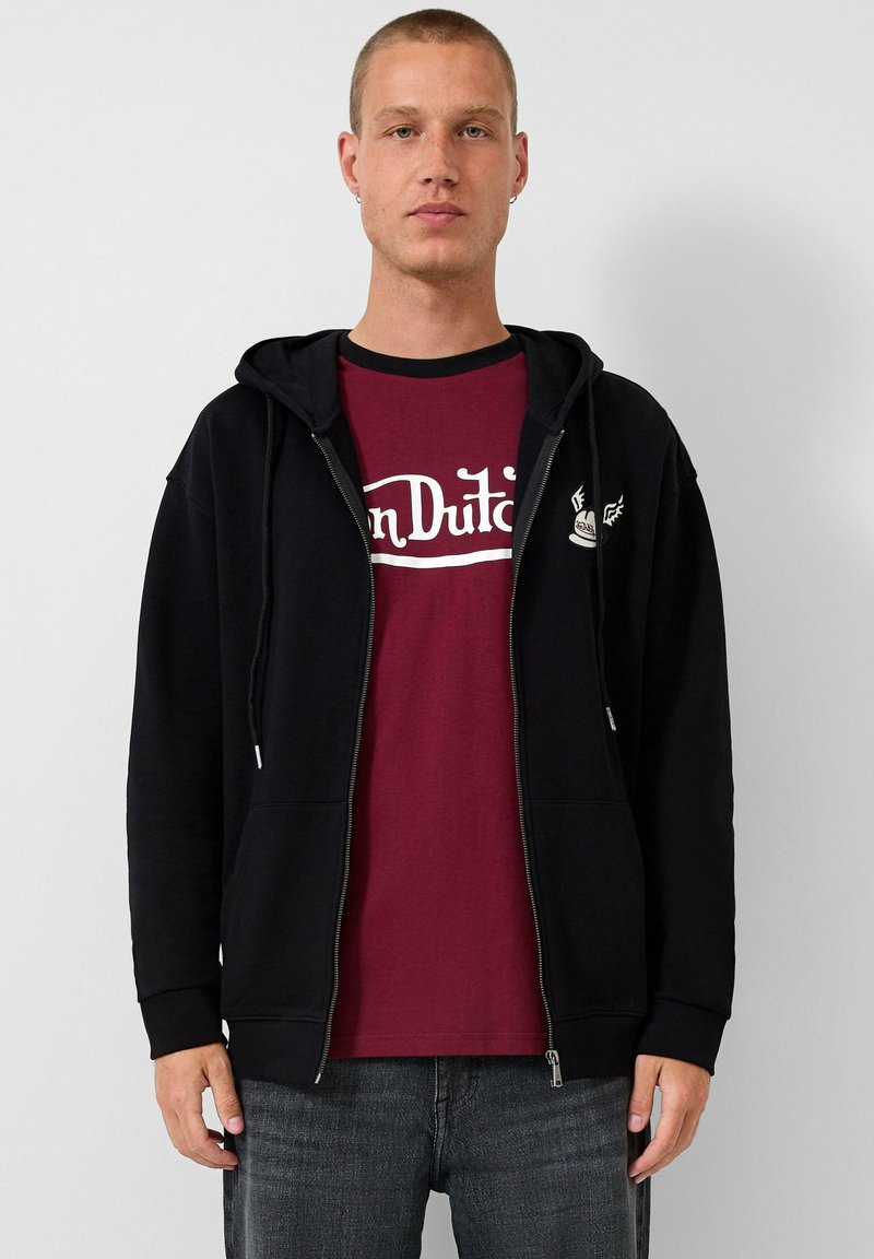 Schwarzer Zip-Hoodie mit Kängurutasche, getragen über einem burgunderfarbenen T-Shirt mit weißem Aufdruck. Das Modell trägt außerdem graue Jeans.