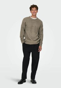 Suéter de color marrón claro, con textura acanalada y cuello redondo, llevado sobre una camiseta blanca, combinado con pantalones negros de corte holgado y zapatos negros.