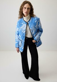 Ajurasti plavi kaput s bijelim cvjetnim uzorcima i crnim obrubom, uparen s crnim širokim hlačama i kremastom puloverom s visokim ovratnikom.