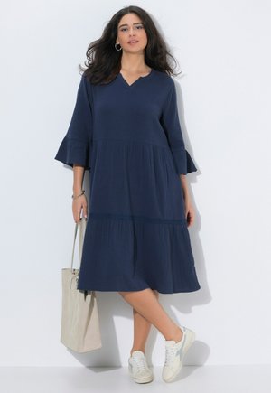 Žena v tmavě modrých midi šatech s rozšířenými rukávy, v bílých teniskách, s kruhovými náušnicemi, držící béžovou tašku před bílou zdí.