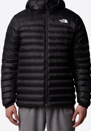 Veste matelassée noire à capuche avec fermeture éclair frontale et logo blanc The North Face sur la poitrine gauche, portée avec un pantalon de survêtement gris.