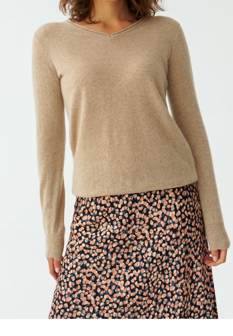 Maglione beige lavorato a maglia con scollatura a V, maniche lunghe e polsini a coste abbinato a una gonna nera con un motivo floreale rosa e beige.