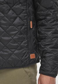 Veste matelassée noire avec extérieur en nylon texturé, fermeture éclair latérale avec tirette en cuir et étiquette de la marque à l'ourlet.