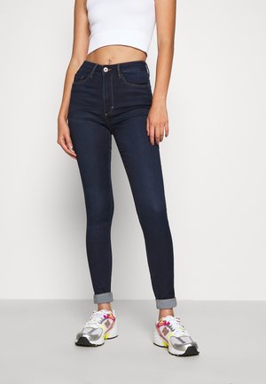 Tummansinisiä denimmitkäisiä skinny-farkkuja, joissa on korkea vyötärö, etumaiset nappikiinnitykset ja käännetyt lahkeet. Yhdistetty valkoiseen lyhytpaitaiseen toppiin ja monivärisiin lenkkareihin.