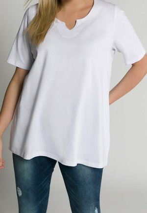 Frau, die ein schlichtes weißes T-Shirt mit V-Ausschnitt und kurze Ärmel sowie blaue, abgenutzte Jeans trägt, steht mit einer Hand in der Hüfte vor neutralem Hintergrund.