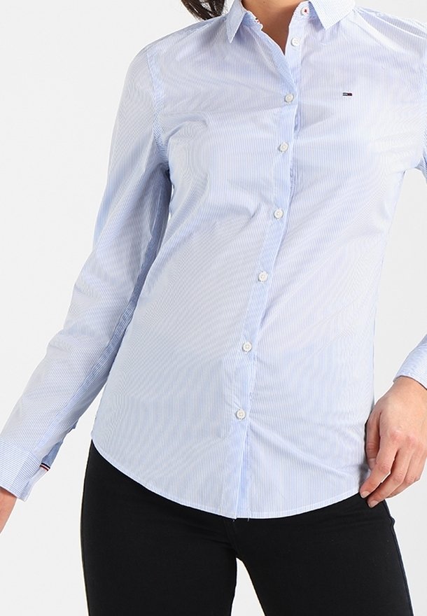 Femme portant une chemise ajustée à rayures bleu clair avec boutons, manches longues et pantalon noir.