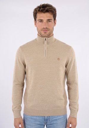 Uomo con maglione beige a mezza zip e jeans azzurri chiari, in piedi davanti a uno sfondo chiaro e uniforme, volto verso avanti con espressione neutra.