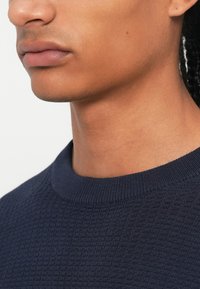 Pull en maille texturée bleu marine avec un col rond, présentant un subtil motif tissé et un col côtelé, présenté sur fond blanc.