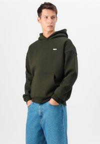Olijfgroene hoodie met een kenguruzak en geribde manchetten, voorzien van een klein wit "OBEY" logo op de borst, gecombineerd met blauwe jeans.