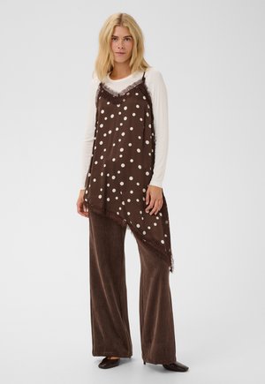 Femme en robe nuisette asymétrique à pois marron superposée sur un haut blanc à manches longues et un pantalon marron à jambes larges, debout face à l'avant.