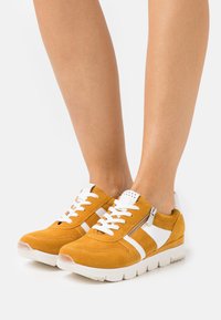 Yellow suede sneakers with white accents, lace-up front, side zipper, and thick white sole. Features perforated detailing on the upper.