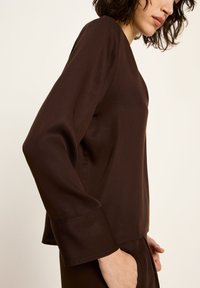 Frau in einem dunkelbraunen Langarmoberteil und passender Hose, mit einer Hand in der Tasche, von der Seite vor einem schlichten hellen Hintergrund gezeigt.