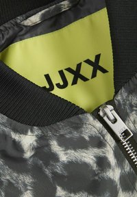 Interno di una giacca bomber nera e grigia con un motivo testurizzato, caratterizzato da un rivestimento giallo brillante e una cerniera argentata con il nome del marchio "JJXX."