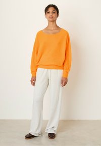Orange oversized sweater med vid halsudskæring, parret med løse lys grå sweatpants og brune slip-on sko, mod en neutral baggrund.