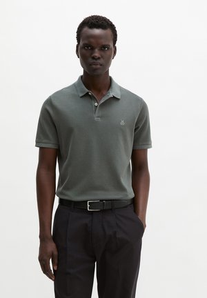 Homme portant un polo à manches courtes vert olive avec un petit logo brodé, ceinture noire et pantalon noir devant un fond blanc uni.
