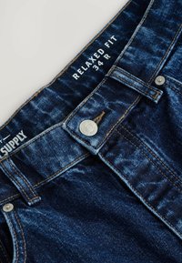 Jeans de mezclilla con un corte relajado, lavado oscuro y costuras claras. Presenta un botón de metal y etiquetas que indican la talla 34 R y la marca.