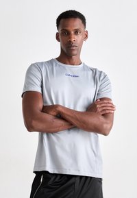 Tricou sportiv cu mânecă scurtă de culoare gri deschis, cu un finisaj texturat și logo-ul albastru "Calvin Klein" pe piept, asortat cu pantaloni scurți negri.