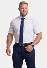 Blonder Mann in weißem Kurzarmhemd, marineblauer Krawatte und dunkler Hose lächelt, schaut nach rechts, linke Hand nahe am Gürtel und trägt eine Armbanduhr.