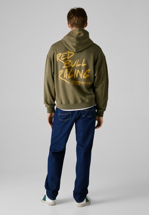 Olive green hoodie with "RED BULL RACING" in bold gold lettering on the back, paired with dark blue jeans and white sneakers with green accents.