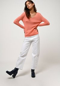 Pfirsichfarbener V-Ausschnitt-Pullover, weiche Textur, kombiniert mit weißen Slim-Fit-Jeans und navyfarbenen Blockabsatz-Stiefeln. Einfaches, modernes Design.