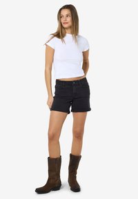 Balta prigludusi marškinėliai, juodi džinsiniai šortai su sukneštais kraštais, rudai aukšti batai iki kelių, stovinti poza su rankomis ant liemenio, neutraliame fone.