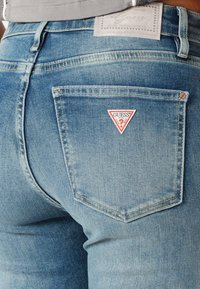 Vaaleansiniset farkut, joissa on kolmioruutuinen punavalkoinen "Guess"-logo takataskussa. Kangas on haalistunut ja siinä on näkyvät saumat.