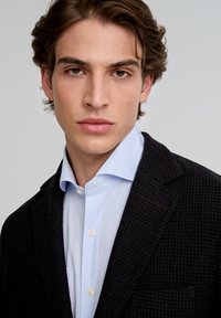 Blazer de pata de gallo oscura sobre una camisa a rayas azul claro, que cuenta con un cuello clásico, cierre frontal de botones y un sutil detalle en el bolsillo.