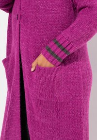 Lange cardigan in fel magenta breiwerk, met een knoopsluiting, zijzakken en donkere gestreepte accenten op de manchetten. Zachte textuur.