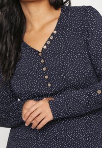 Námořnické šaty s dlouhým rukávem, které mají bílé puntíky, klopu a dřevěné knoflíky vpředu. Měkká tkanina s hladkým povrchem.