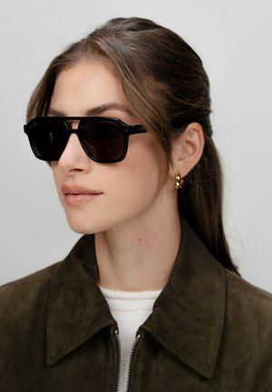 Occhiali da sole neri rettangolari con lenti scure, caratterizzati da una montatura spessa e bordi angolari. Il modello indossa una giacca in suede verde scuro e orecchini a cerchio dorati.