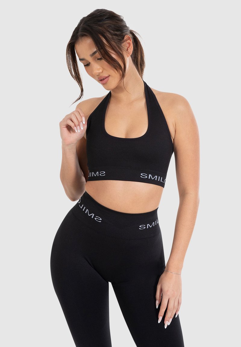 Zwarte halternek sportbh met een vierkante halslijn; gecombineerd met hoge zwarte leggings met een witte logo band in de taille.