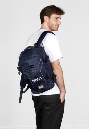 Mochila azul marino con el logo de la NASA y un parche de la bandera estadounidense, con un diseño estructurado, correas ajustables y múltiples orificios de ventilación.