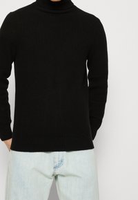 Suéter negro de cuello alto con canalé, de mangas largas y diseño ajustado, combinado con vaqueros azul claro. Textura suave, con detalles mínimos.