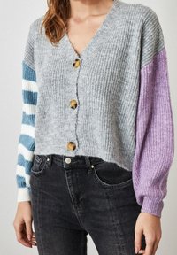 Graue, gerippte Strickjacke mit V-Ausschnitt, versehen mit drei großen goldenen Knöpfen und mehrfarbigen gestreiften Ärmeln in Blau- und Lilatönen.