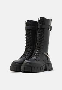 Schwarze knöchelhohe Plattformstiefel mit dicken Gummisohlen, Schnürung vorne, seitlichem Reißverschluss und einer Metallakzentkette an der Seite. Glattes Lederfinish.