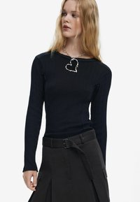 Fekete textúrájú, hosszú ujjú felső, kerek nyakkivágással, sötét szoknyával párosítva, amely övvel és oldalsó zsebekkel rendelkezik. Ezüst szívnyaklánc díszíti.