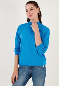 Sudadera azul de manga larga, con cuello alto, puños acanalados y un corte relajado. Combinada con jeans azules que muestran una textura suave.