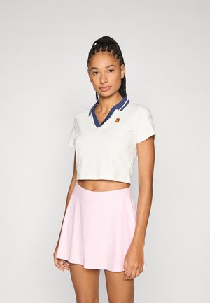 Polo blanc court avec col bleu marine, texture à motifs et petit logo. Associé à une jupe évasée rose clair, tissu lisse.