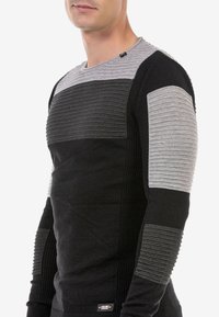 Schwarzer und grauer strukturierten Pullover mit horizontalen Rippstrukturen, langen Ärmeln und schlanker Passform. Enthält ein kleines Etikett auf der unteren linken Seite.