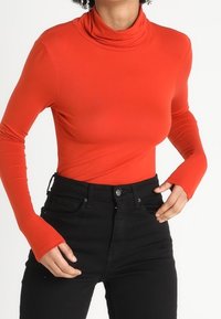 Röd långärmad polotröja i slätt tyg, figurnära design med hög krage, matchad med högmidjade svarta jeans.