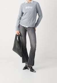 Sweatshirt gris avec le texte "BOSS" blanc, superposé à une chemise blanche à col. Jeans gris foncé évasés avec des ourlets effilochés et des bottines noires à cheville. Sac fourre-tout noir.